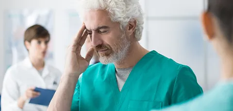 Male doctor in teal scrubs touching his temple, illustrating diagnostic challenge of Bell’s palsy versus stroke facial weakness.